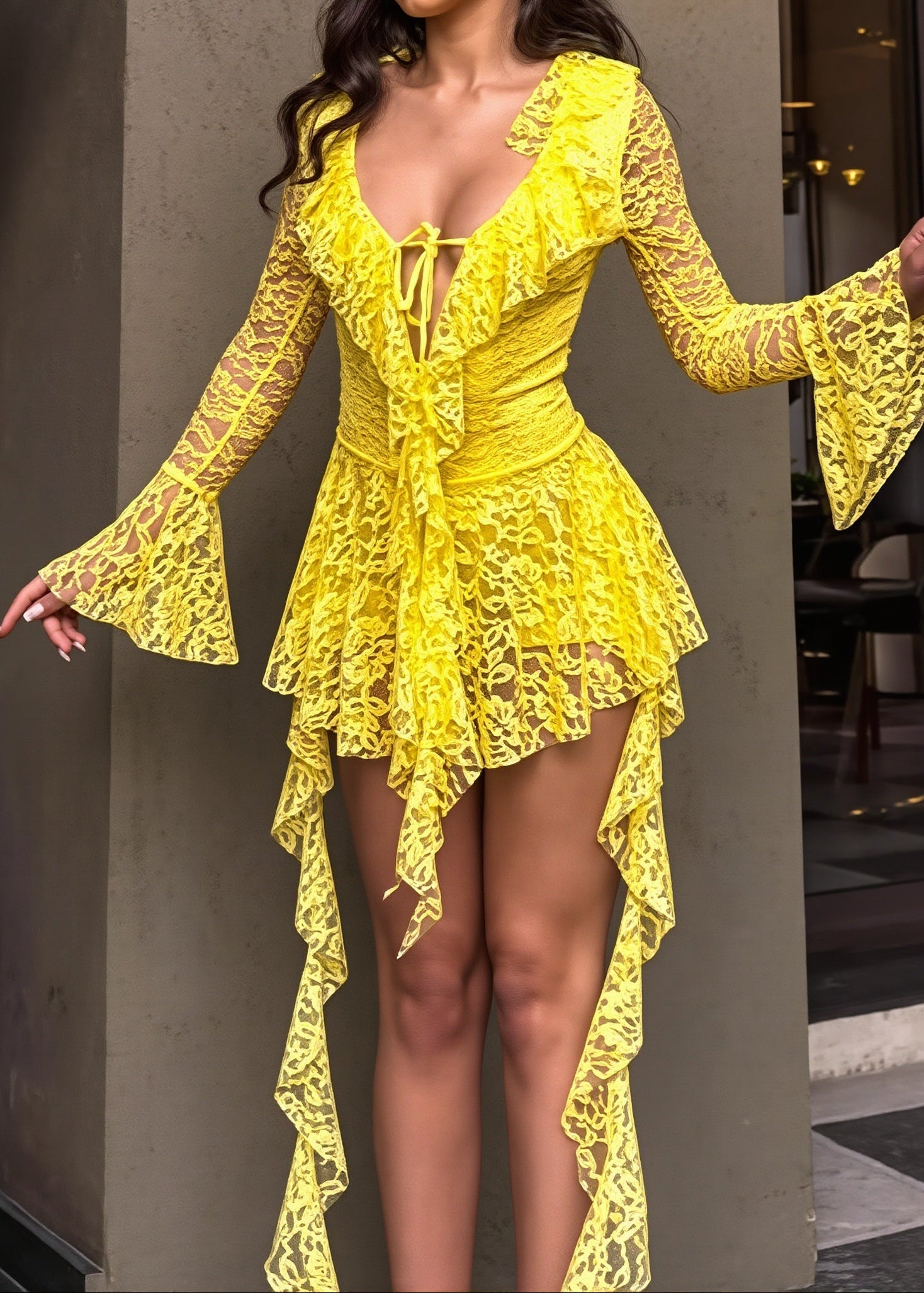 A woman modeling a bright yellow lace dress with long flared sleeves, a deep V-neckline, and an asymmetrical high-low hem, posed against a modern concrete pillar background.