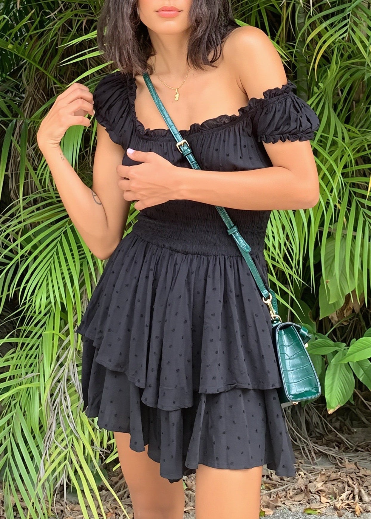 A woman wearing a black off-the-shoulder ruffled mini dress with a tiered skirt, standing outdoors in front of lush green palm foliage and holding a small green shoulder bag.