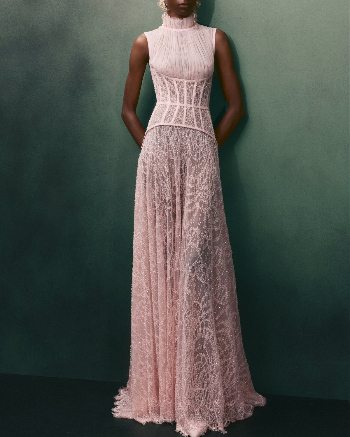 A model stands against a dark green textured wall wearing a sleeveless, high-neck, floor-length pink lace gown with a structured corset-style bodice.