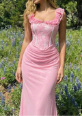 A woman wearing a pink sleeveless maxi dress with a ruffled sweetheart neckline, lace corset-style bodice, and gathered skirt, standing outdoors in a field of blue flowers and green foliage.