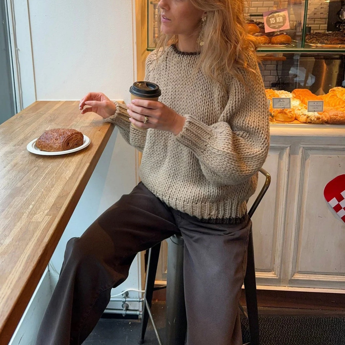 Oatmeal chunky open knit deep V-back oversized sweater - seated side shot in bakery cafe showing relaxed boxy silhouette with dark leather wide-leg trousers