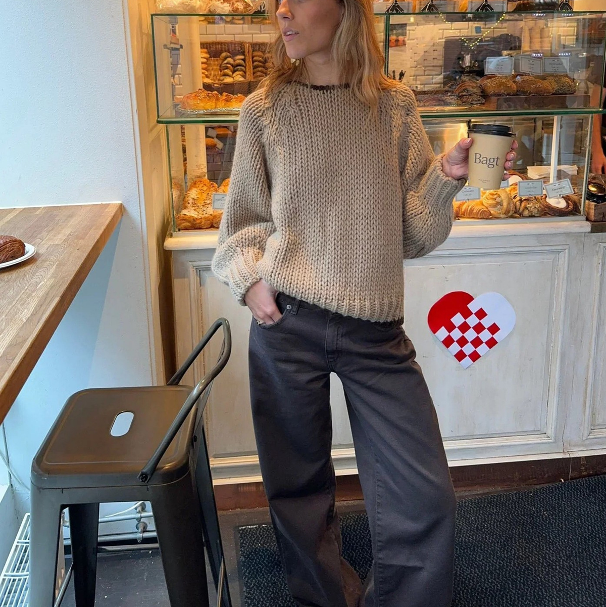 Oatmeal chunky open knit deep V-back oversized sweater - front standing shot in bakery with dark chocolate leather wide-leg trousers and stacked rings holding coffee cup