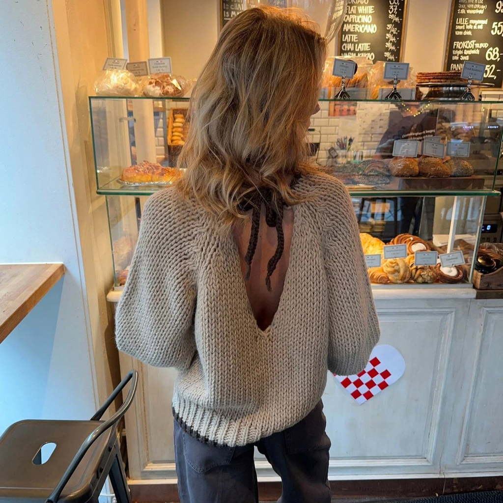 Oatmeal chunky open knit deep V-back oversized sweater - back view in bakery showing dramatic deep V-back with self-tie string detail and open knit texture with dark jeans