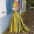 Olive gold satin halter neck open back ball gown maxi dress - outdoor full length editorial showing structured bodice sculptural bow waist and voluminous sweep train with silver glitter clutch