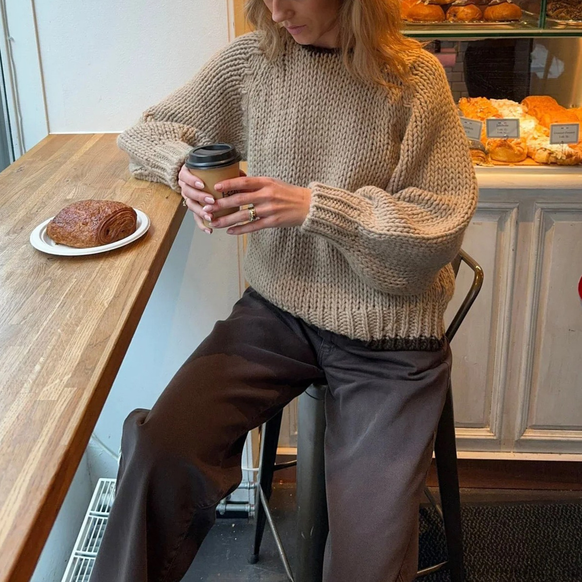 Oatmeal chunky open knit deep V-back oversized sweater - seated three-quarter shot in bakery showing chunky ribbed cuff detail holding coffee cup with stacked rings